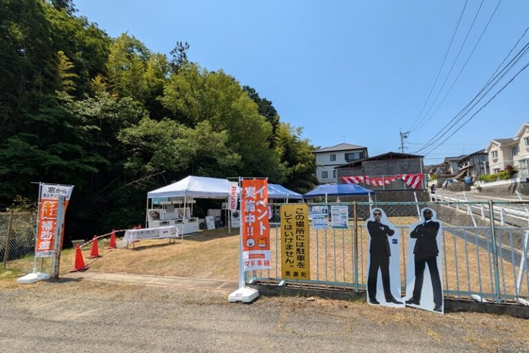 イベントの様子。手前にスーツ姿の外国人のパネル、警備部門SATのイメージキャラクター。 奥に紅白幕があり、テントを張ってガス機器やリフォーム商材、ホームセキュリティなど展示している。